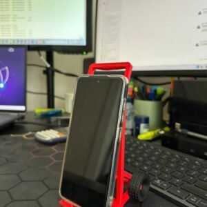 May include: A bright red plastic smartphone stand shaped like a two-wheeled hand truck. The stand features a rectangular frame with a base ledge to support a mobile device and two small black wheels attached to the sides for a realistic aesthetic.