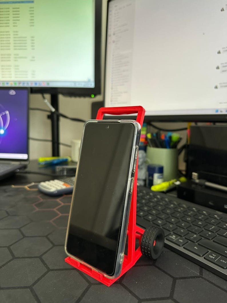 May include: A bright red plastic smartphone stand shaped like a two-wheeled hand truck. The stand features a rectangular frame with a base ledge to support a mobile device and two small black wheels attached to the sides for a realistic aesthetic.