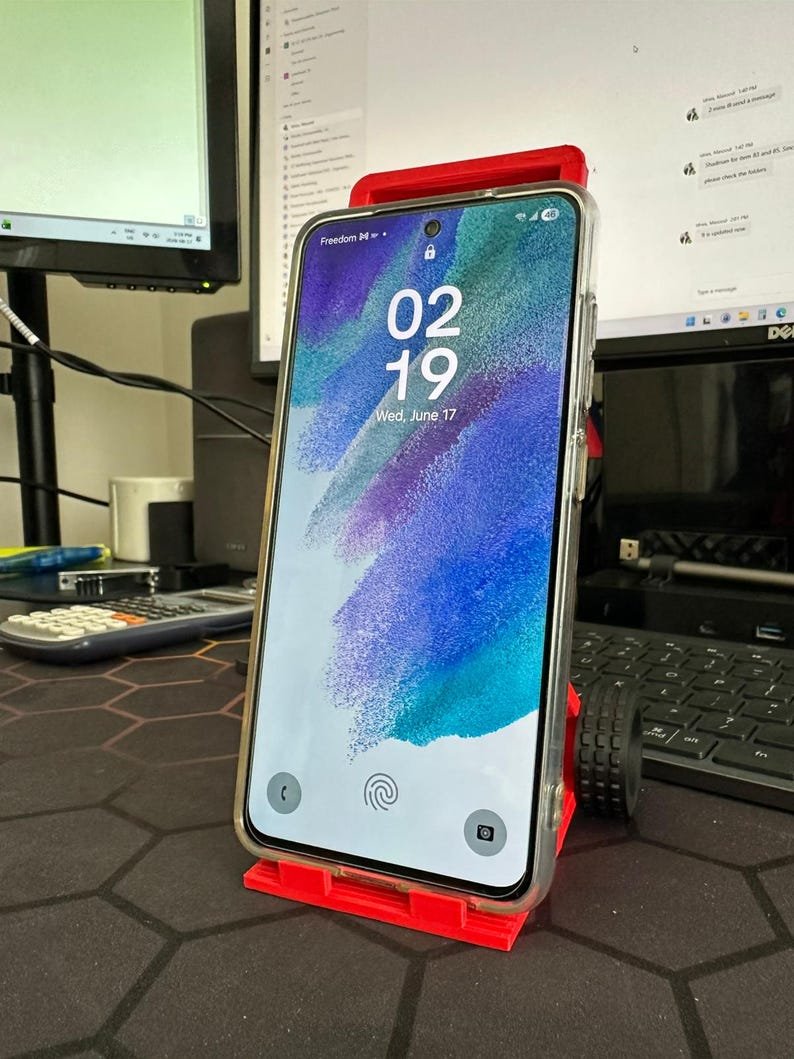 3D Printed Mini Hand Truck Dolly Phone Stand May include: A smartphone with a clear protective case resting on a bright red plastic stand. The screen displays the time 02:19, the date Wed, June 17, and a fingerprint icon. The stand features a small black wheel-like detail on the side.