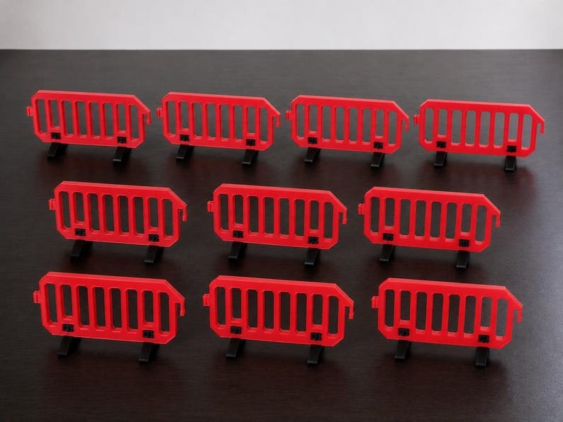 Miniature UK Traffic Cones & Roadwork Barriers Set May include: A set of ten miniature red plastic crowd control barriers, each featuring a rectangular frame with vertical slats and two black feet for stability, arranged in three rows on a dark surface.