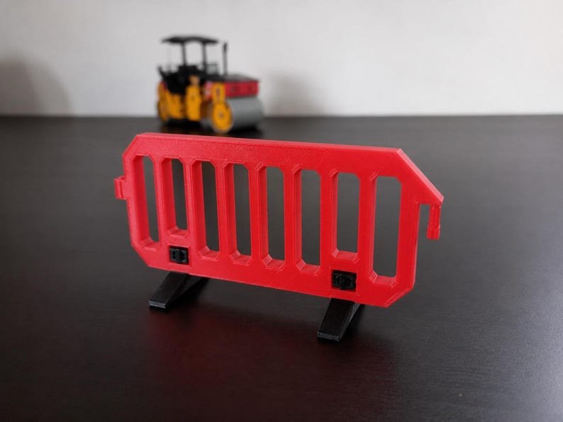 Miniature UK Traffic Cones & Roadwork Barriers Set May include: A miniature red plastic crowd control barrier featuring a series of vertical slats and two black detachable feet. The barrier is positioned in the foreground, while a small yellow and black toy road roller is visible in the background.