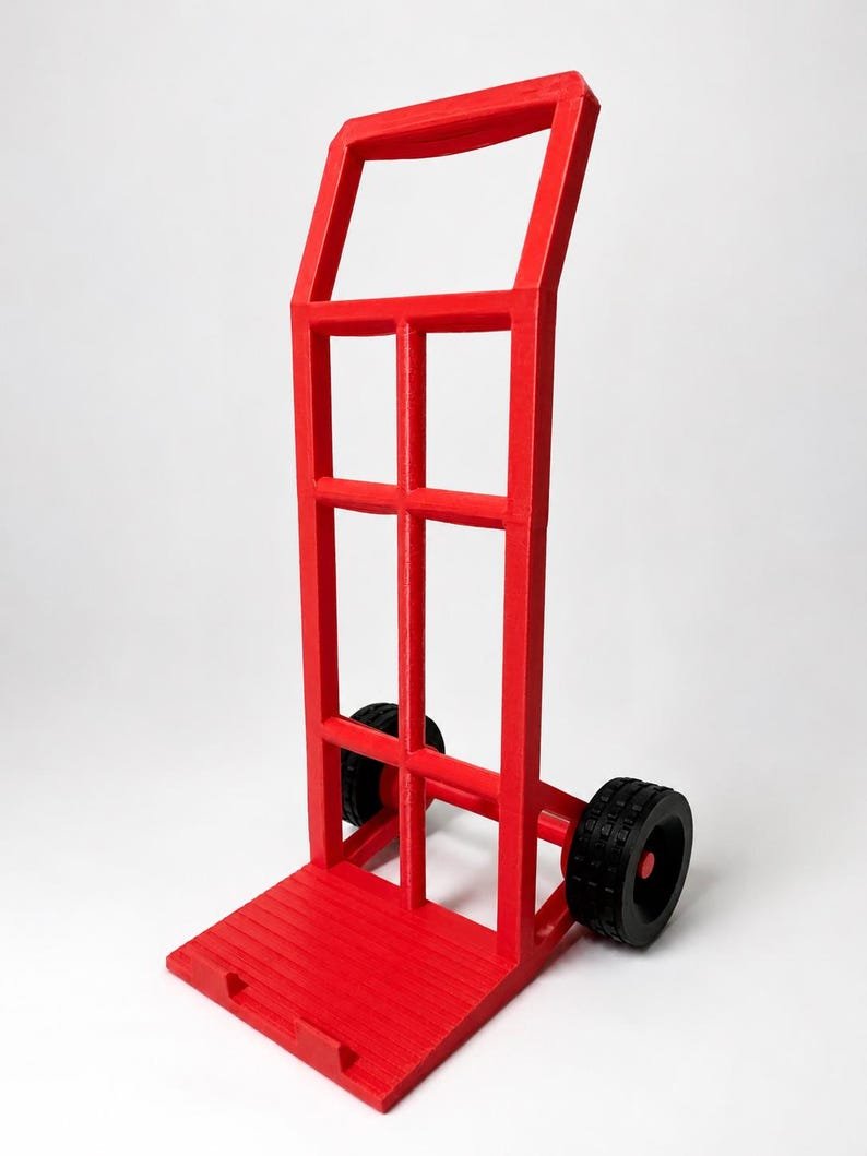3D Printed Mini Hand Truck Dolly Phone Stand May include: A bright red hand truck featuring a tall, rectangular frame with a handle at the top and a flat, grooved base plate. The unit is supported by two black wheels with textured treads, positioned on a horizontal axle at the lower rear section.