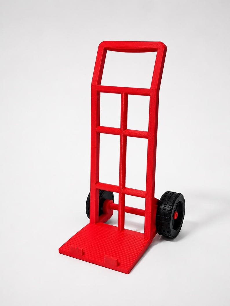 3D Printed Mini Hand Truck Dolly Phone Stand May include: A bright red hand truck featuring a tall, rectangular frame with a grid-like structure, a flat base plate with two small protruding tabs, and two black wheels with a textured tread pattern attached to the lower sides of the frame.