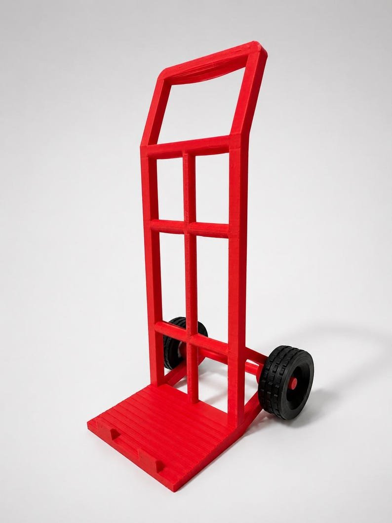 3D Printed Mini Hand Truck Dolly Phone Stand May include: A bright red hand truck featuring a tall, rectangular frame with a handle at the top and a flat base plate at the bottom. The unit is equipped with two black wheels attached to a horizontal axle, providing mobility for the transport device.