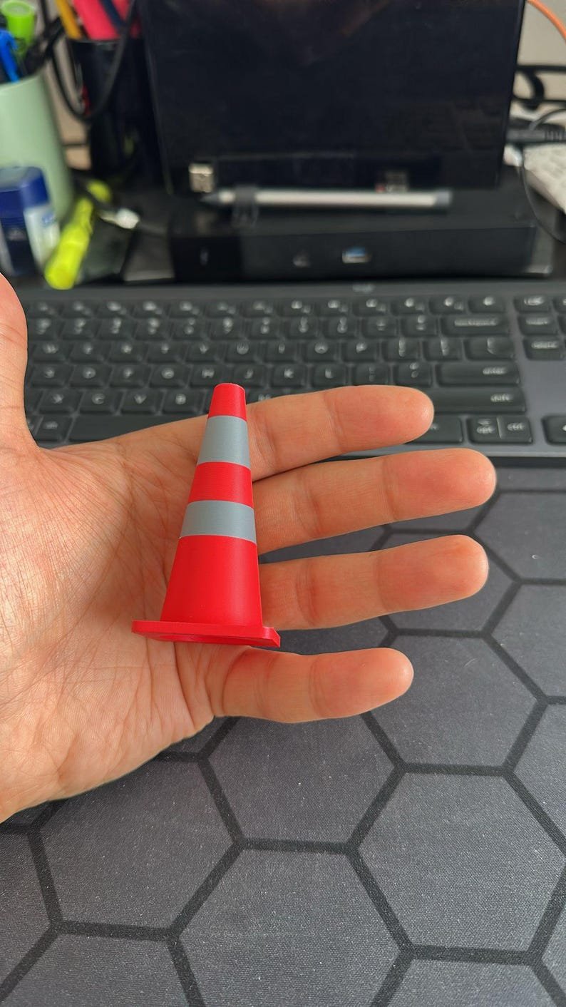 Mini Traffic Cones — 2.76-inch May include: A small, bright red traffic cone featuring two horizontal grey reflective stripes. The cone has a wide, square-shaped base and a smooth, matte finish, designed as a miniature replica of standard road safety equipment.