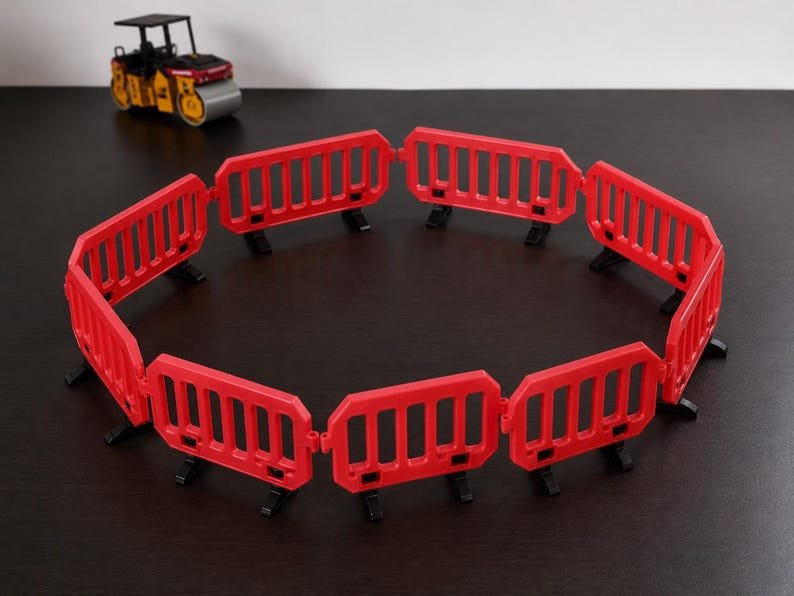 Miniature UK Traffic Cones & Roadwork Barriers Set May include: A circular arrangement of eight red plastic interlocking construction barriers, each featuring vertical slats and black feet, positioned on a dark surface with a small yellow and black toy road roller visible in the background.