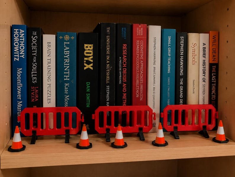 Miniature UK Traffic Cones & Roadwork Barriers Set May include: A row of books on a shelf featuring three red plastic construction barriers and five miniature orange and white traffic cones placed in front of the book spines. The books include titles like Moonflower Murders, Labyrinth, and Boy X.