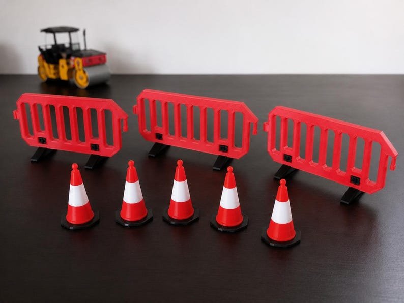 Miniature UK Traffic Cones & Roadwork Barriers Set May include: A set of three red plastic construction barriers with vertical slats and black feet, accompanied by five red and white traffic cones. In the background, a small yellow and black toy road roller is visible on the dark surface.