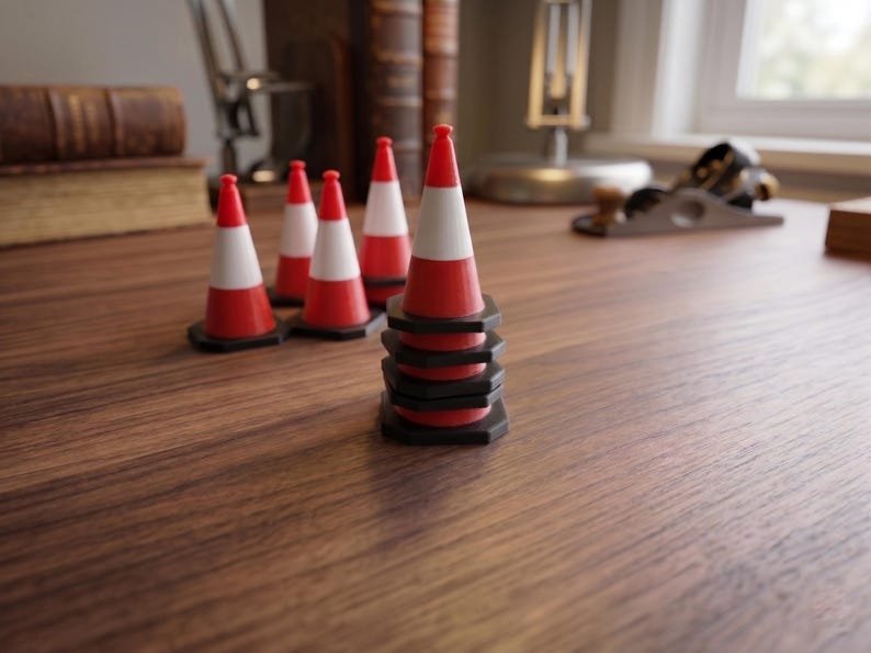 Miniature UK Traffic Cones & Roadwork Barriers Set May include: A collection of miniature traffic cones featuring a red and white striped design with a black hexagonal base. One cone stands prominently in the foreground, while four additional cones are arranged in a cluster behind it.