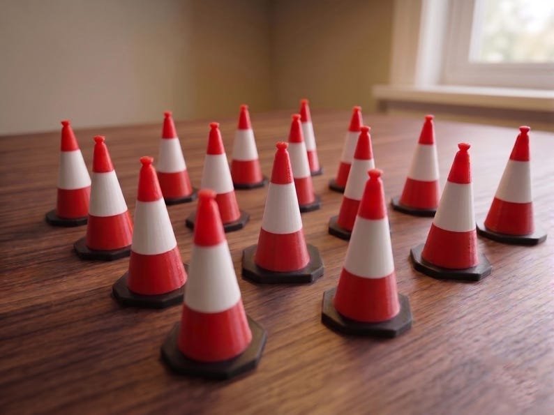 Miniature UK Traffic Cones & Roadwork Barriers Set May include: A collection of fifteen miniature traffic cones arranged in a group. Each cone features a bright orange top and base with a distinct white horizontal stripe around the middle, all mounted on a black octagonal base for stability.