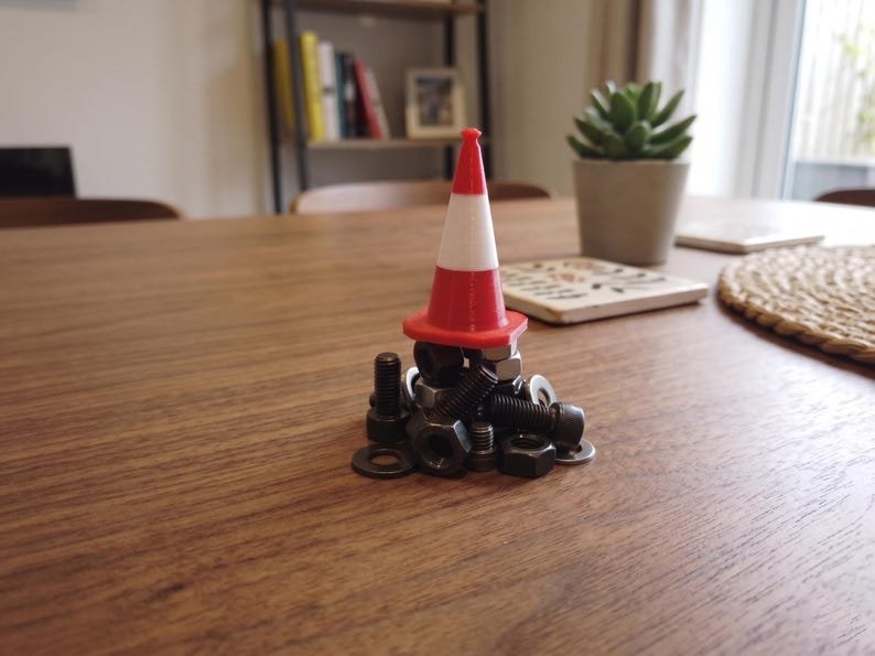 Miniature UK Traffic Cones & Roadwork Barriers Set May include: A small red and white plastic traffic cone sits atop a pile of assorted black metal hardware, including several hexagonal nuts, bolts, and flat washers. The cone features a distinct horizontal white stripe around its centre.