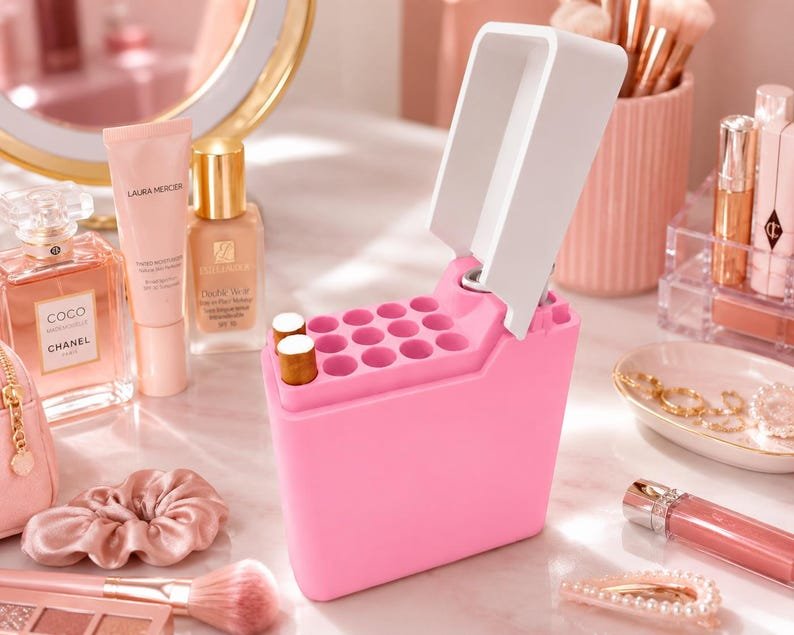 Pink Cigarette Case with Lighter Holder — King Size, 15 Cigarettes May include: A bright pink makeup organiser with a hinged white lid and multiple circular slots for holding lipsticks or brushes. The organiser stands upright among various cosmetic bottles, a makeup brush, a scrunchie, and jewellery items.