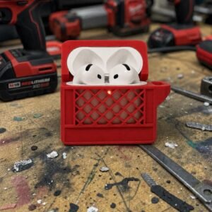 May include: A pair of white wireless earbuds housed inside a red protective case designed to resemble a plastic milk crate. The case features a grid-like exterior pattern and a small circular cutout on the front, revealing a glowing orange indicator light.