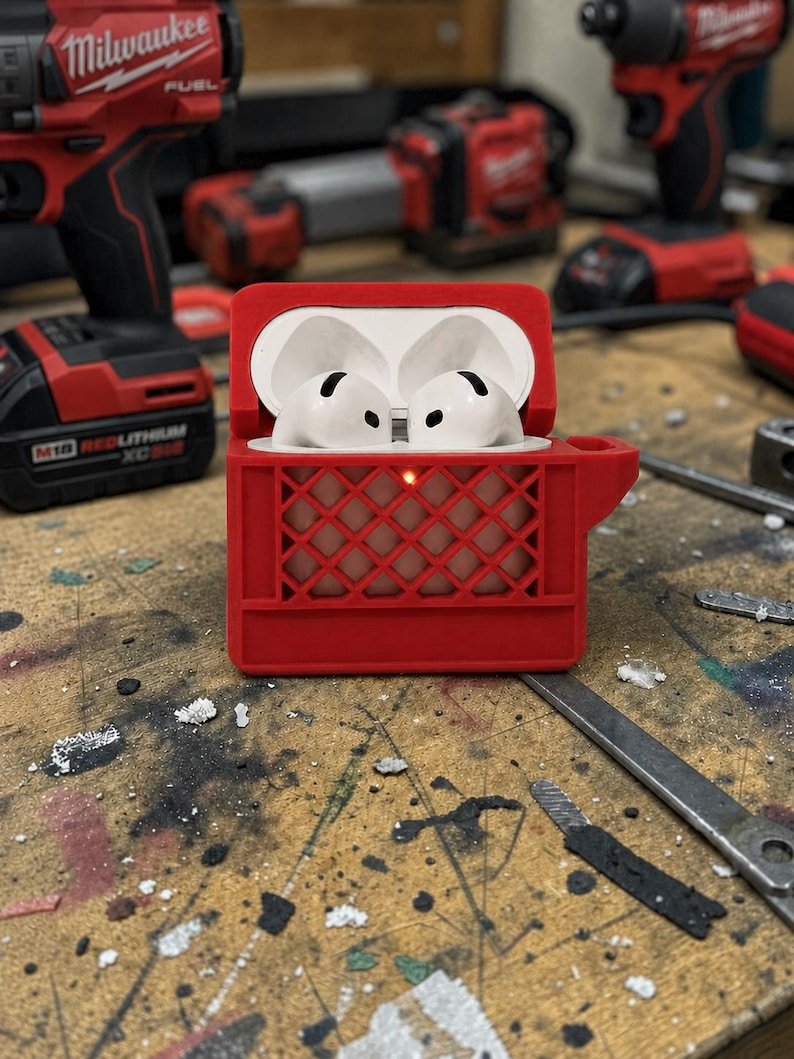 Mini Milk Crate AirPods 4 Case May include: A pair of white wireless earbuds housed inside a red protective case designed to resemble a plastic milk crate. The case features a grid-like exterior pattern and a small circular cutout on the front, revealing a glowing orange indicator light.
