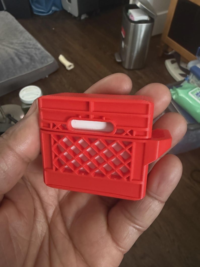 Mini Milk Crate AirPods 4 Case May include: A bright red protective case for wireless earbuds, designed to resemble a miniature plastic milk crate. The case features a rigid, grid-like structure with a rectangular handle cutout on the top section and a diamond-patterned side.