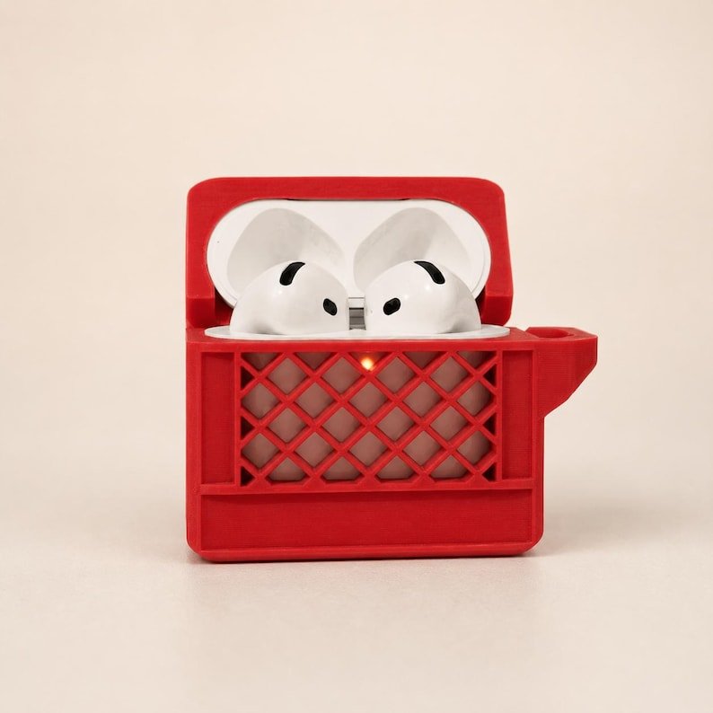 Mini Milk Crate AirPods 4 Case May include: A bright red protective case for wireless earbuds, designed to resemble a miniature milk crate with a lattice pattern. The case is open, revealing two white earbuds inside, and features a small cutout on the side for a charging port.