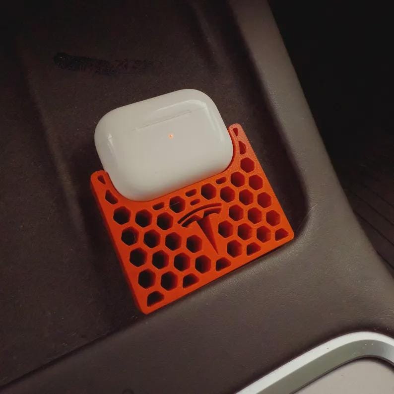 AirPods Pro 2/3 Wireless Charging Spacer for Tesla May include: A white wireless earbud charging case sits inside an orange, rectangular holder featuring a honeycomb lattice pattern and a Tesla logo embossed in the centre. The holder is designed to fit securely within a vehicle console area.