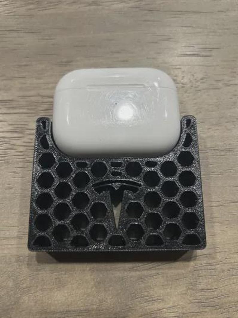 AirPods Pro 2/3 Wireless Charging Spacer for Tesla May include: A white wireless earbud charging case rests inside a black, 3D-printed holder. The holder features a textured, honeycomb-patterned exterior and displays a prominent Tesla logo embossed in the centre of the front panel.
