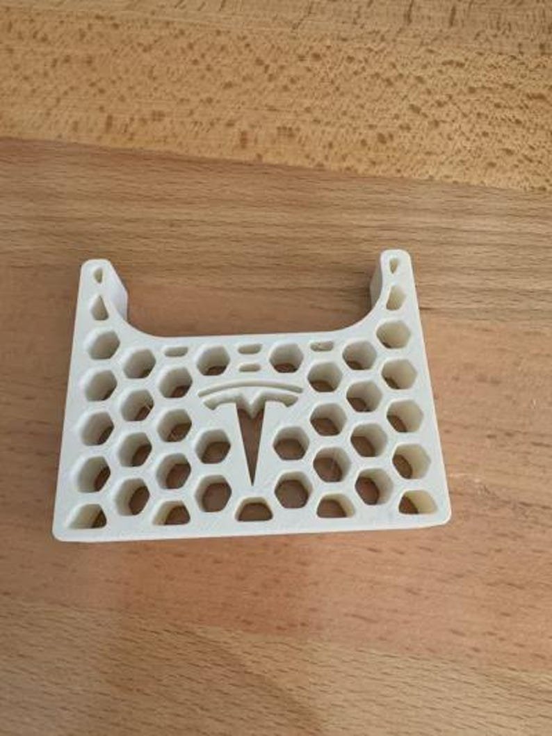 AirPods Pro 2/3 Wireless Charging Spacer for Tesla May include: A white, 3D-printed plastic accessory featuring a repeating hexagonal honeycomb pattern throughout its structure. The centre of the piece displays a prominent Tesla logo cutout, and the top edge has a curved, U-shaped indentation.