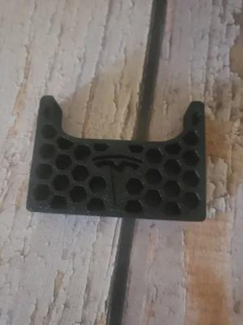AirPods Pro 2/3 Wireless Charging Spacer for Tesla May include: A black, rectangular, 3D-printed plastic component featuring a honeycomb grid pattern throughout its surface. Centered within the hexagonal grid is a raised Tesla logo. The top edge has a U-shaped cutout, and two small circular protrusions are visible on the upper corners.