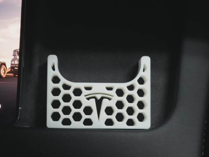 AirPods Pro 2/3 Wireless Charging Spacer for Tesla May include: A white, rectangular, 3D-printed storage insert featuring a honeycomb pattern and a central Tesla logo cutout. The object has a curved top edge and is designed to fit into a vehicle console, providing a structured space for small items.