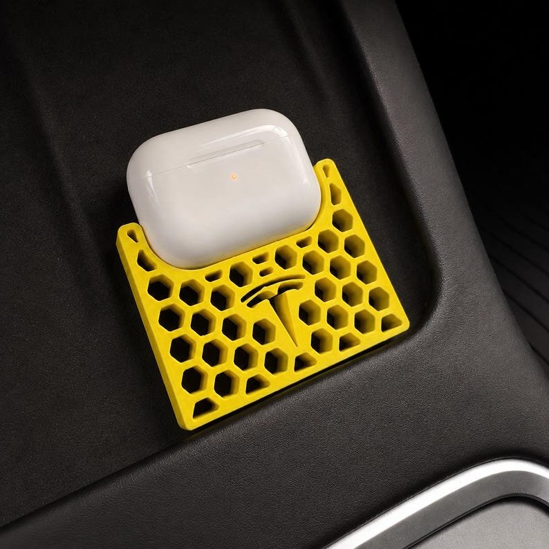 AirPods Pro 2/3 Wireless Charging Spacer for Tesla May include: A bright yellow, honeycomb-patterned holder featuring a Tesla logo cutout, designed to securely store a white wireless earbud charging case. The holder has a geometric, open-cell structure and is positioned on a dark, smooth surface.