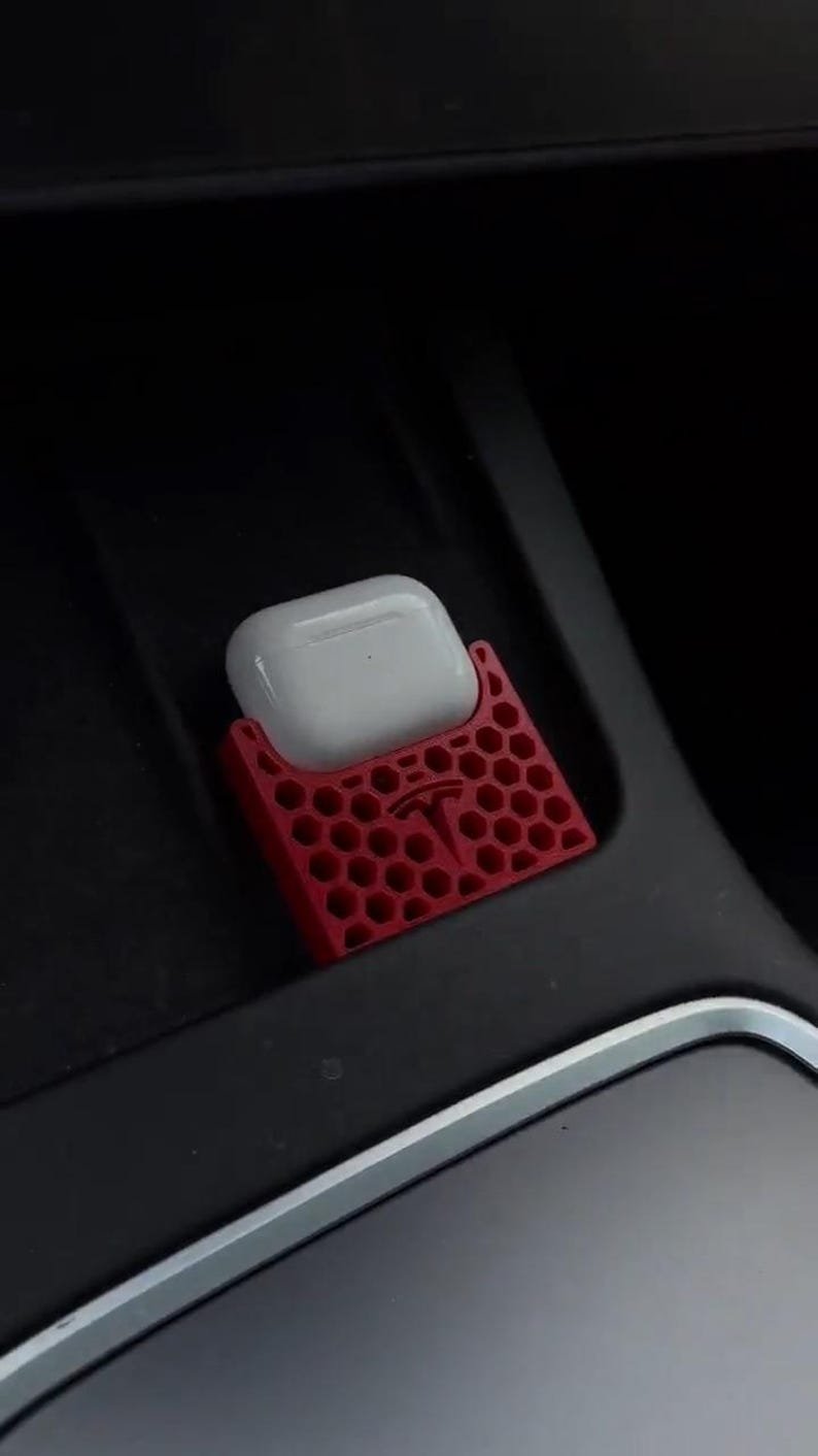 AirPods Pro 2/3 Wireless Charging Spacer for Tesla May include: A white wireless earbud charging case sits inside a red, 3D-printed plastic holder with a honeycomb pattern and a Tesla logo embossed on the front. The holder is designed to fit into a specific slot within a vehicle's centre console.
