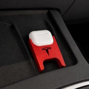 May include: A white wireless earphone charging case sits inside a red plastic holder featuring the Tesla logo. The holder is designed with a cutout base and is positioned within a dark grey textured storage compartment of a vehicle interior.