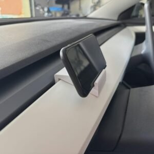 May include: A white plastic smartphone mount secured to the dashboard of a vehicle. The mount holds a black smartphone in a vertical position, angled towards the driver. The dashboard features a white trim piece and a dark grey textured surface.