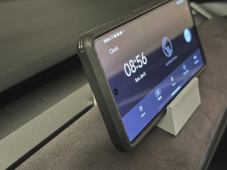 Tesla Model 3/Y Phone Holder May include: A smartphone in a black protective case resting on a light grey angled stand. The screen displays a digital clock showing 08:56, the date Sun, Jan 5, and various app icons including Alarm, Clock, Timer, and World Clock in a dark blue interface.