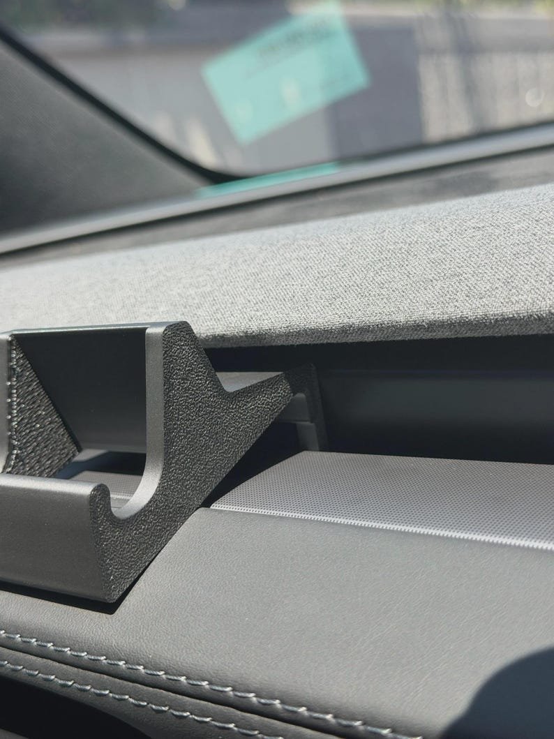 Tesla Model 3/Y Phone Holder May include: A black, textured plastic mount with a Y-shaped cradle design, positioned on a vehicle dashboard. The mount features a rigid, angular structure with a recessed centre, designed to securely hold an object against the car interior.