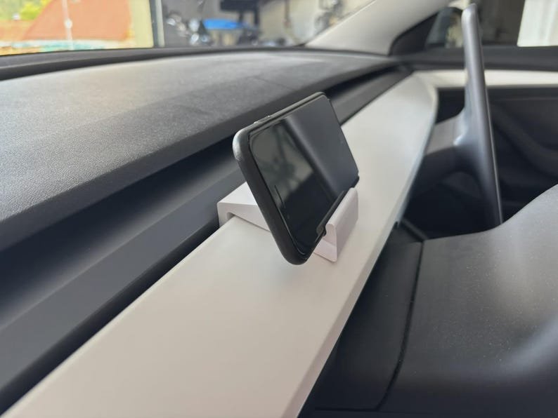 Tesla Model 3/Y Phone Holder May include: A white plastic smartphone mount secured to the dashboard of a vehicle. The mount holds a black smartphone in a vertical position, angled towards the driver. The dashboard features a white trim piece and a dark grey textured surface.