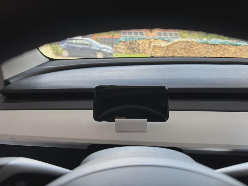 Tesla Model 3/Y Phone Holder May include: A black smartphone mounted in a white rectangular stand positioned on a flat surface. The device is oriented horizontally, displaying a dark screen. The stand is solid and provides a stable base for the phone within a vehicle interior.