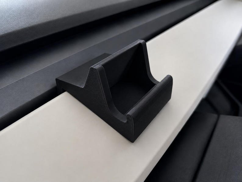Tesla Model 3/Y Phone Holder May include: A matte black, 3D-printed plastic mount with a wedge-shaped base and a curved cradle designed to hook onto a flat, horizontal surface. The object features a textured finish and a hollow centre, providing a stable support structure.