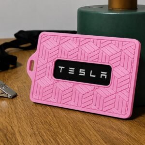 May include: A rectangular pink silicone badge holder featuring a geometric embossed pattern and a central black window displaying the white text TESLA. The holder includes a side slot for a lanyard attachment and is paired with a black lanyard clip.