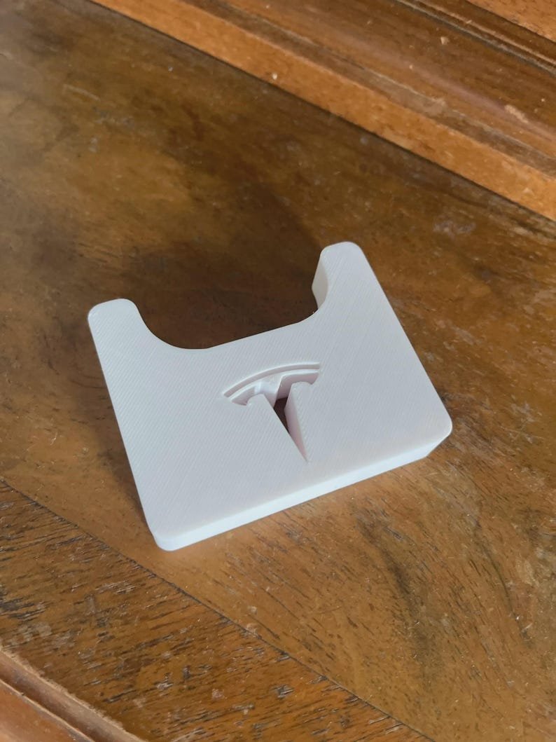 Tesla Model 3 & Model Y Airpods 4 Charging Stand Spacer May include: A white, 3D-printed plastic holder featuring a cutout of the Tesla logo in the centre. The object has a rectangular base with a curved indentation at the top edge, designed for mounting or storage purposes with a smooth, matte finish.