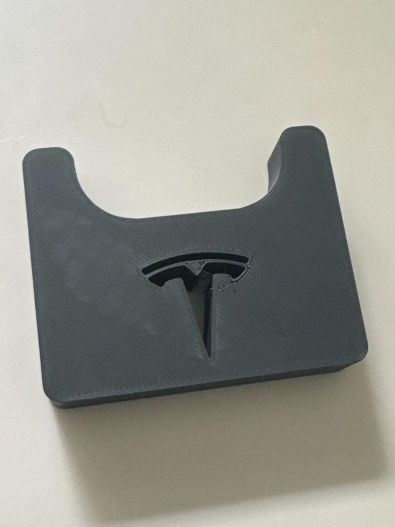 Tesla Model 3 & Model Y Airpods 4 Charging Stand Spacer May include: A dark grey, rectangular, 3D-printed object featuring a U-shaped cutout at the top edge and a recessed Tesla logo centred on the front surface. The item has a matte finish with visible layer lines from the manufacturing process.