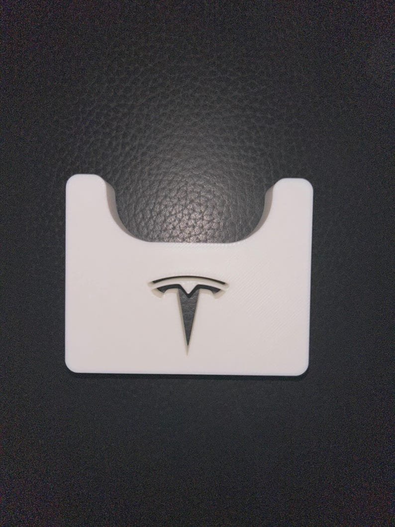 Tesla Model 3 & Model Y Airpods 4 Charging Stand Spacer May include: A white rectangular plastic holder featuring a cutout at the top centre and a black Tesla logo embossed in the middle. The object has a smooth finish and a flat base, designed with a distinct U-shaped indentation along the top edge.