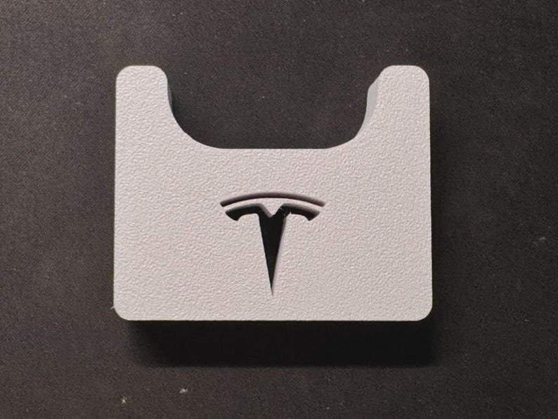 Tesla Model 3 & Model Y Airpods 4 Charging Stand Spacer May include: A light grey, rectangular, textured block featuring a U-shaped cutout at the top centre and a black Tesla logo embossed in the middle. The object has a flat base and rounded corners, presenting a solid, uniform appearance throughout.