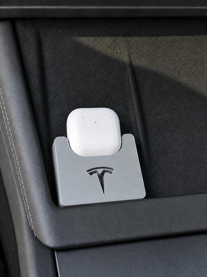 Tesla Model 3 & Model Y Airpods 4 Charging Stand Spacer May include: A white wireless earbud charging case sits inside a grey, rectangular holder featuring a cutout of the Tesla logo. The holder is positioned on a dark, textured surface, and the charging case displays a small, illuminated orange light.