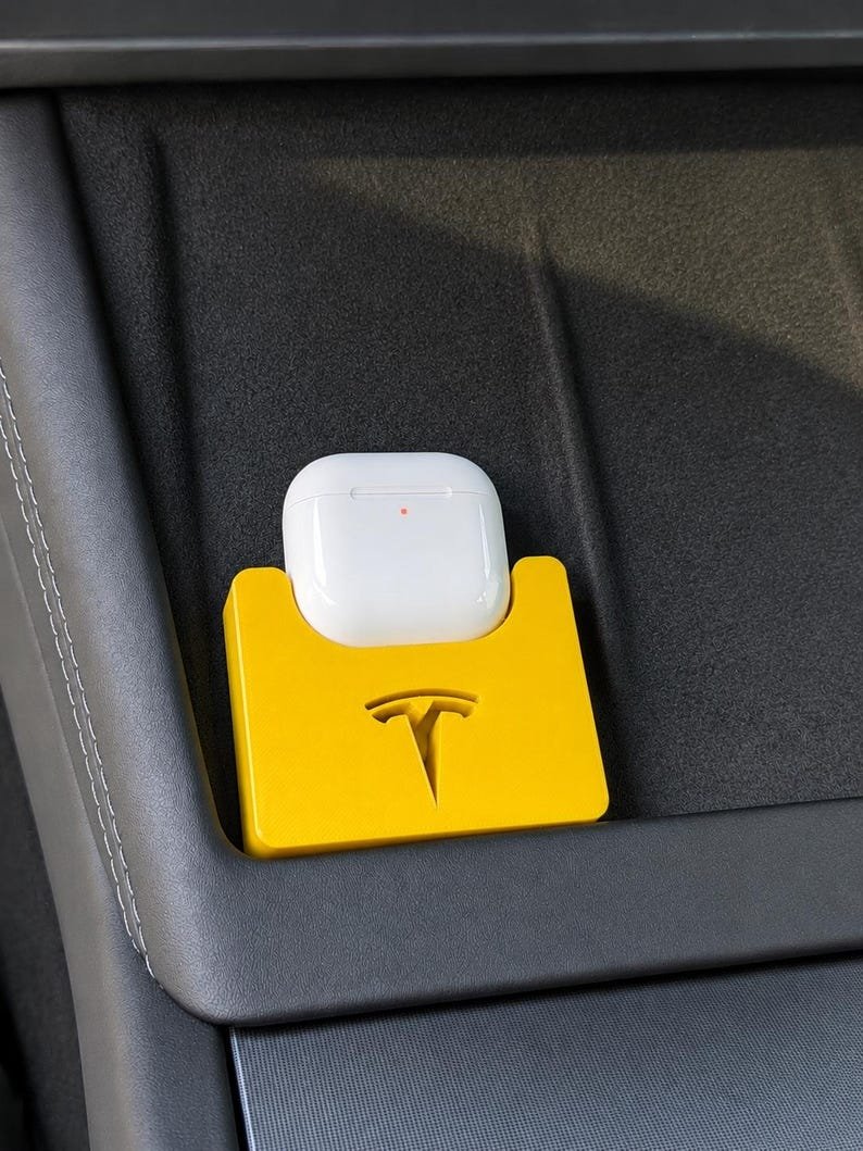 Tesla Model 3 & Model Y Airpods 4 Charging Stand Spacer May include: A bright yellow rectangular holder featuring a cutout of the Tesla logo. The holder is designed to securely store a white wireless earbud charging case, which is currently placed inside the slot with a small orange indicator light visible.
