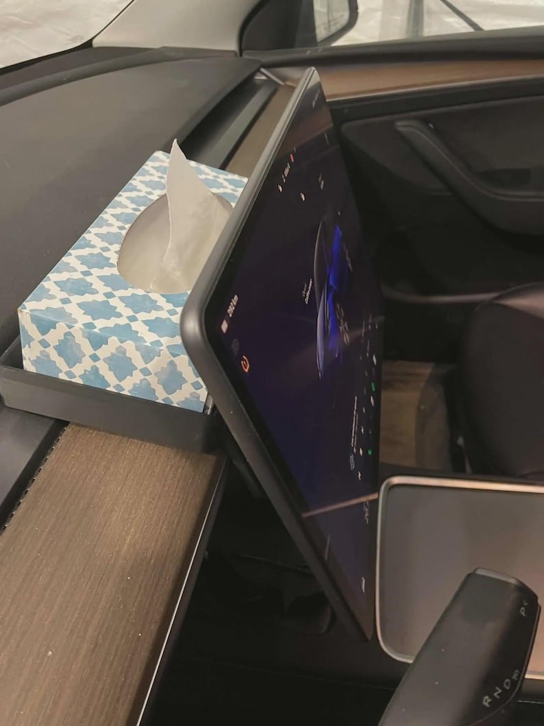 Tesla Model 3 & Model Y Tissue Box Holder May include: A rectangular tissue box with a blue and white geometric pattern sits on the dashboard of a vehicle next to a large, vertically oriented black touchscreen display. The screen shows a digital interface with a vehicle graphic and icons.