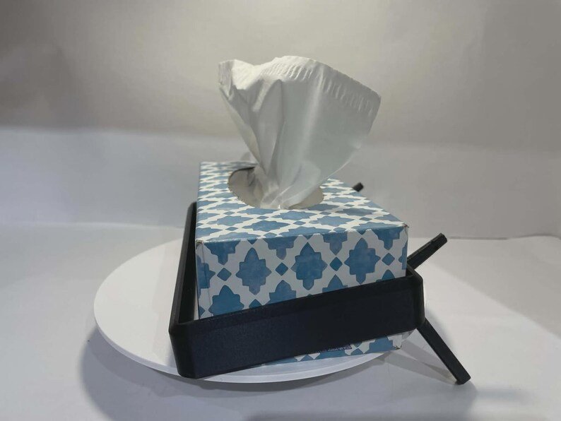 Tesla Model 3 & Model Y Tissue Box Holder May include: A rectangular tissue box with a blue and white geometric pattern, containing white tissues, held within a black metal frame stand. The stand features angled supports on the sides, providing a stable base for the tissue box container.