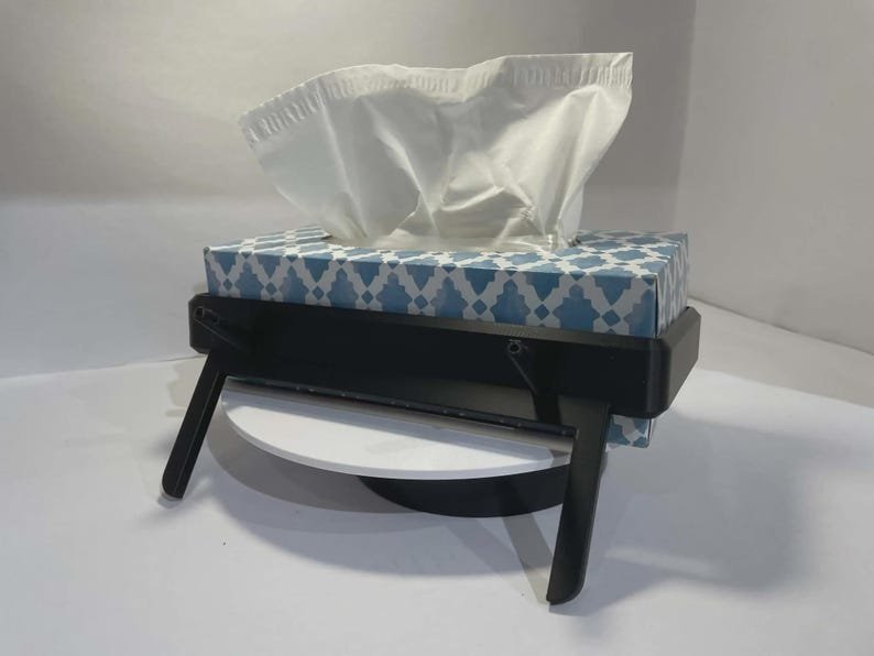 Tesla Model 3 & Model Y Tissue Box Holder May include: A rectangular tissue box with a blue and white geometric pattern, containing white tissues, held within a black plastic stand featuring two angled legs that elevate the box off the surface for a tilted display position.