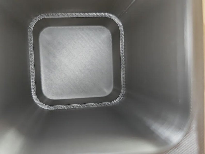 Mini Wheelie Bin Trash Can Pencil Holder May include: A top-down view of a square, dark grey container with rounded corners. The interior walls show a visible, fine-textured, layered pattern consistent with 3D printing, creating a smooth, uniform finish across the base and sides.