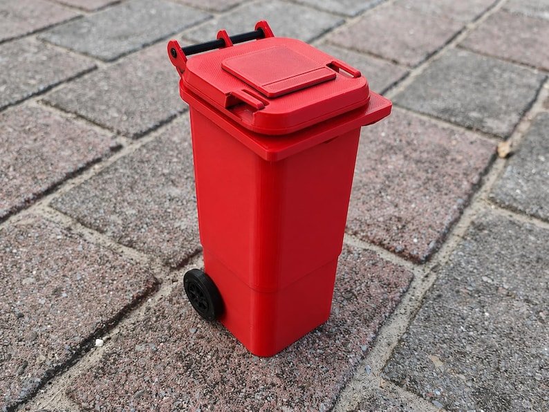 Mini Wheelie Bin Trash Can Pencil Holder May include: A miniature red wheelie bin featuring a hinged lid and two black wheels. The bin has a rectangular body, a flat top cover with a small handle, and a black axle connecting the wheels at the base, designed as a small-scale replica.