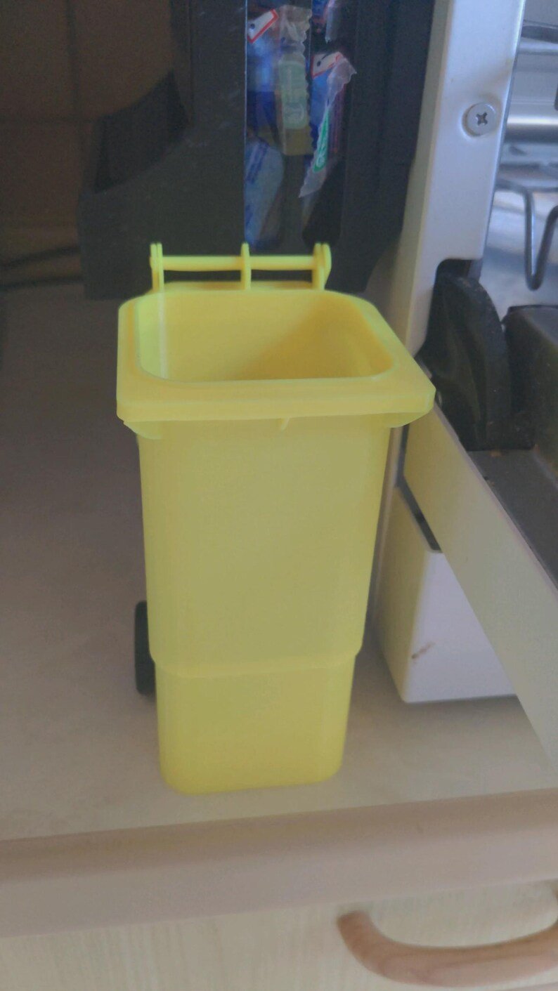 Mini Wheelie Bin Trash Can Pencil Holder May include: A small, bright yellow plastic waste bin featuring a rectangular body, an open top, and a hinged lid mechanism at the back. The container is designed to resemble a standard outdoor refuse bin, complete with a moulded rim and side wheels.
