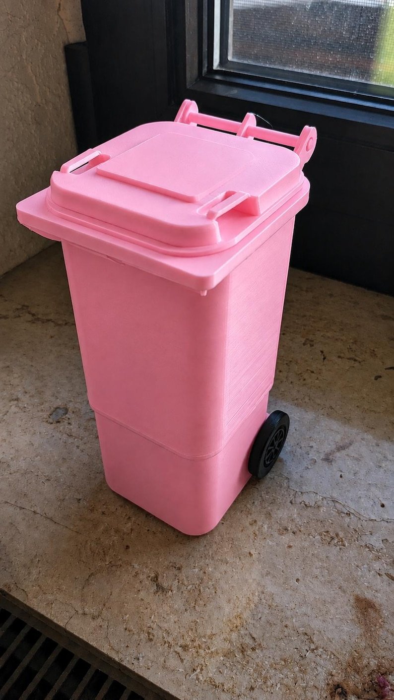 Mini Wheelie Bin Trash Can Pencil Holder May include: A small, bright pink plastic waste bin featuring a hinged lid and two black wheels on the sides. The container has a rectangular body with a flat top and a textured surface, designed to mimic the appearance of a standard outdoor dustbin.
