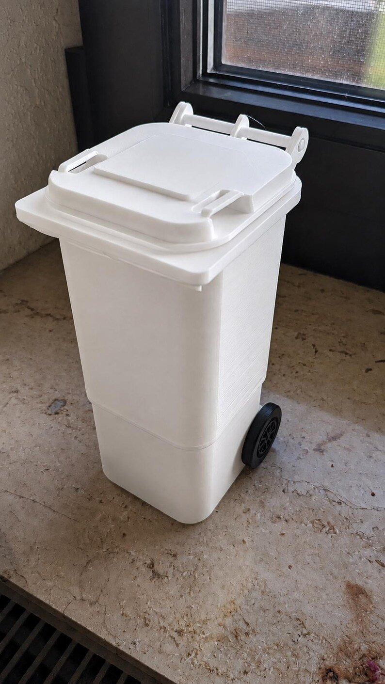 Mini Wheelie Bin Trash Can Pencil Holder May include: A white plastic waste bin featuring a rectangular body, a hinged lid with a handle, and two black wheels attached to the lower sides. The container has a smooth finish and a functional design that mimics a standard outdoor refuse bin.