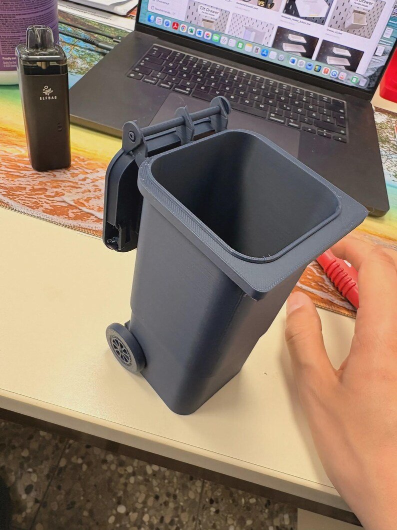 Mini Wheelie Bin Trash Can Pencil Holder May include: A small, dark grey plastic waste bin with a hinged lid and two wheels on the back. The container features a rectangular opening at the top and a textured surface finish, designed to resemble a standard outdoor refuse bin.